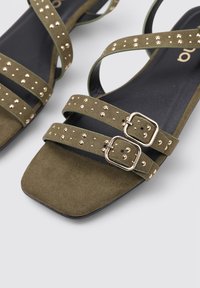 Sandalias de ante verde oliva con correas entrecruzadas, adornadas con tachuelas doradas y hebillas. Diseño de punta cuadrada con suela plana negra.