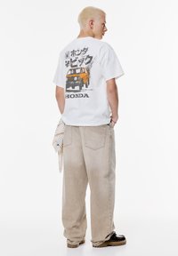 Hvid t-shirt med orange bilgrafik og "HONDA" tekst på ryggen, sammen med lysebeige løstsiddende jeans og sorte sko.