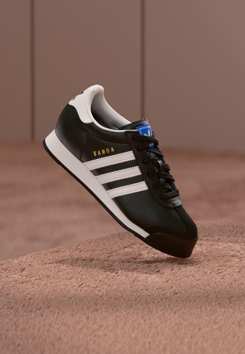 Fekete Adidas Samoa sportcipő fehér csíkokkal és sarokfüllel, arany "Samoa" felirattal, kék logós nyelvvel, barna texturált felület felett lebegve.