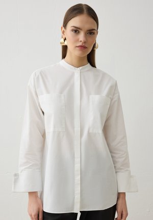 Jeune femme portant une chemise blanche à manches longues boutonnée avec deux poches poitrine et de grandes boucles d'oreilles dorées sur un fond uni.
