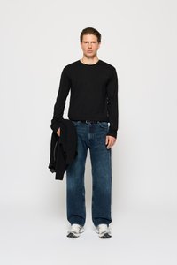 Joven vestido con una camisa negra de manga larga y jeans azules, sosteniendo un suéter negro, de pie contra un fondo blanco liso.