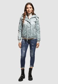 Chaqueta puffer azul claro con un diseño ajustado, cintura con cordón y capucha; combinada con vaqueros azul claros desgastados y botas de tobillo negras brillantes.