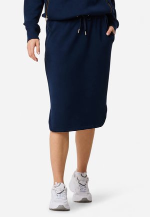 Femme portant une jupe midi bleu marine et des baskets blanches, une main dans la poche et l'autre détendue le long du corps.