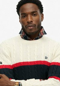 Maglione color crema con trama a trecce, caratterizzato da strisce orizzontali rosse e blu navy. Dettaglio del colletto a quadri molto evidente.