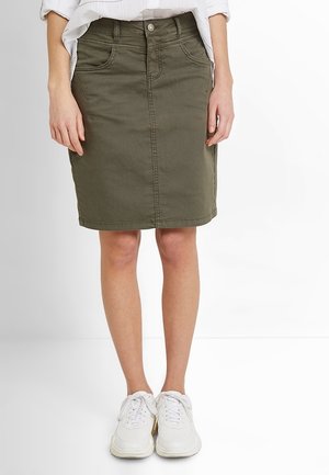 Kobieta w oliwkowo-zielonej, dżinsowej spódnicy do kolan i białych trampkach, stojąca z rękami swobodnie opuszczonymi wzdłuż ciała.