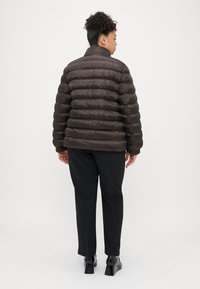 Rjava napihljiva jakna z visokim ovratnikom, prešita zasnova in sijoča tekstura, oblečena čez črne hlače in črne čevlje s peto, pogled od zadaj.