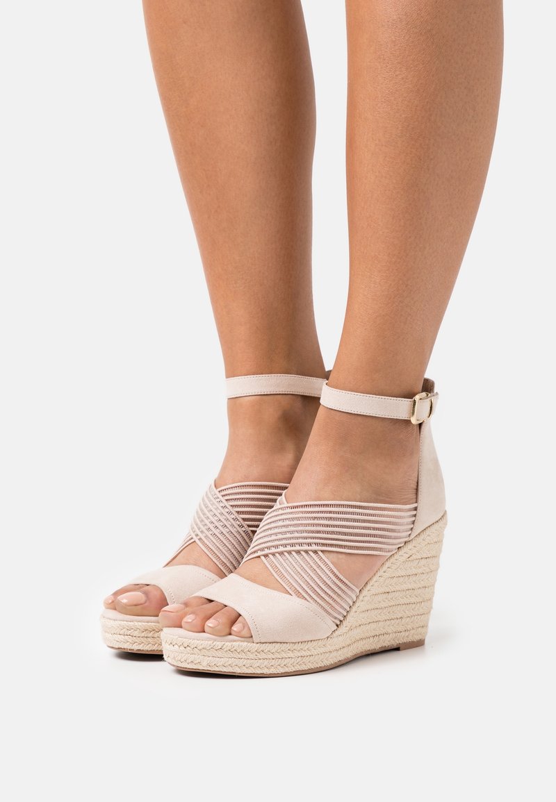 Sandales compensées beige avec brides à la cheville et bandes élastiques croisées portées pieds nus sur un fond uni.