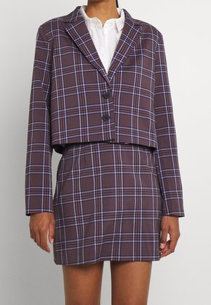 Kvinde iført en kort brun og lilla ternet blazer med sorte knapper over en hvid skjorte med krave og matchende ternet nederdel.