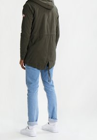Olive green parka jacket with a hood, drawstring waist, and patch detail. Worn over light blue jeans and white sneakers. Plain background.