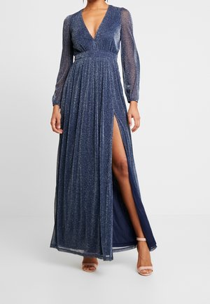 Vrouw draagt een lange, glinsterende donkerblauwe jurk met doorschijnende lange mouwen en een hoge split aan de dij, gecombineerd met lichtroze open-teen hakken.