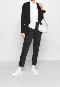 Veste noire à ouverture devant, chemise blanche à boutons, jeans noirs, baskets blanches et un petit sac à main en relief croco blanc avec une bandoulière.