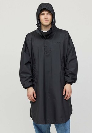 Man draagt een zwarte regenponcho met capuchon en elastische manchetten, staand tegen een effen lichte achtergrond.