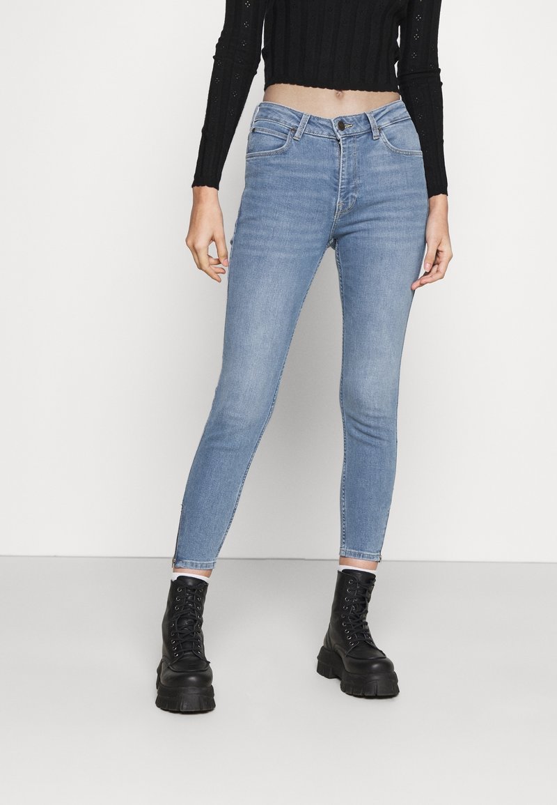 Mujer vistiendo jeans ajustados azul claro, suéter negro corto y botas gruesas negras con cordones, de pie contra un fondo blanco liso.