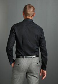 Herren-Fit Schwarzhemd mit langen Ärmeln, kombiniert mit grauen Hahnentritt-Hosen, versehen mit einem glänzenden schwarzen Gürtel und dezenten Manschettendetails.