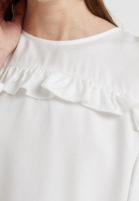Blusa bianca con colletto arricciato. Il tessuto è liscio con una leggera trama, evidenziando un design pulito e minimalista.