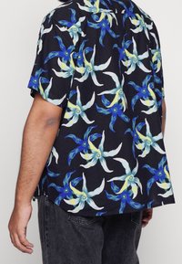 Chemise à manches courtes en tissu noir ornée d'un motif floral bleu et jaune. Coupe décontractée avec un ourlet courbe et un détail texturé subtil.