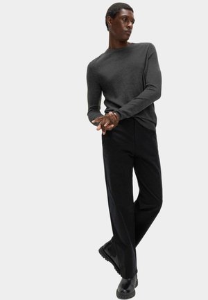 Stående person iført en mørkegrå langærmet sweater, sorte bukser og sorte sko, hænderne foldet, ser til siden på en hvid baggrund.