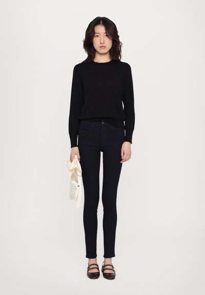 Pulover negru, blugi skinny închis la culoare și pantofi plate maro. Modelul ține o geantă de culoare deschisă. Material neted, design mulat, stil minimalist.