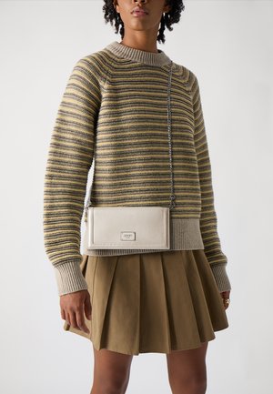 Suéter a rayas en tonos tierra, combinado con una falda de plisada en color caqui. Un pequeño bolso rectangular con herrajes plateados cuelga de una correa de cadena.