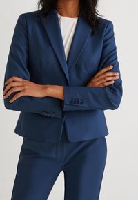 Blazer bleu marine en tissu lisse, avec une coupe ajustée, un design simple boutonnage et trois boutons décoratifs sur les poignets.