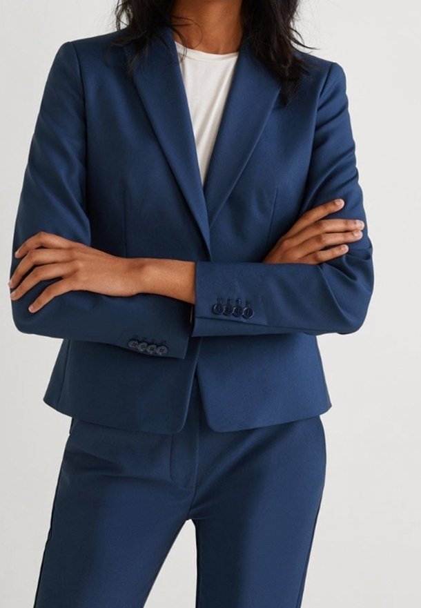 Blazer bleu marine en tissu lisse, avec une coupe ajustée, un design simple boutonnage et trois boutons décoratifs sur les poignets.