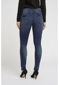 Jean skinny en denim bleu foncé, avec une taille mi-haute, deux poches arrière et un léger dégradé. Associé à des chaussures à talons hauts noires.