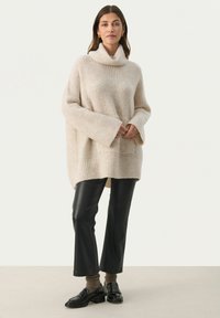 Beige strikket sweater med høj krave, brede ærmer og ribbet tekstur, parret med sorte læderbukser og kraftige sorte loafers.