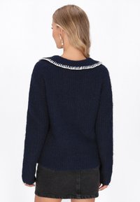 Granatowy sweter z teksturowanej tkaniny, z białą falbanką przy kołnierzyku, długimi rękawami i luźnym krojem. Widok z tyłu pokazuje dół kończący się na wysokości talii.