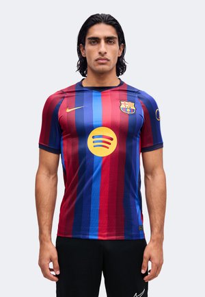 Homme aux cheveux noirs mi-longs portant un maillot de football rayé rouge et bleu du FC Barcelone avec des logos de sponsors, debout devant un fond clair.