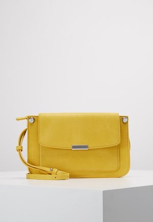 Sac bandoulière en cuir jaune avec rabat avant et fermoir rectangulaire argenté, reposant sur un socle blanc sur fond uni.