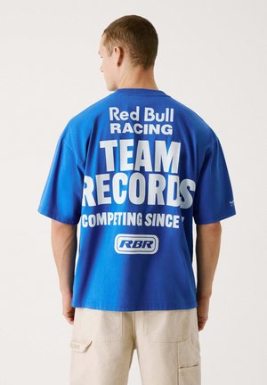 Mėlyna medvilnė marškinėliai su didelėmis baltomis raidėmis ant nugaros: "Red Bull Racing KOMANDOS ĮRAŠAI VARŽOSI NUO" su logotipu apačioje.