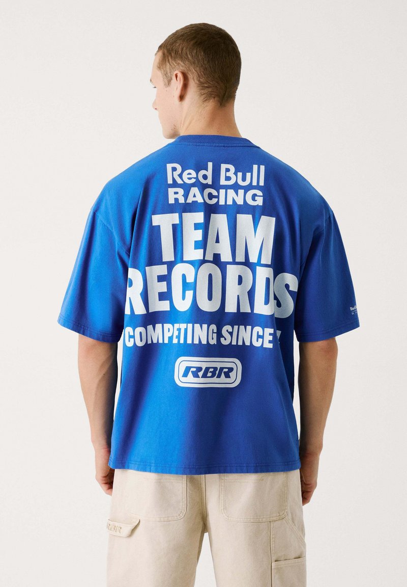 Mėlyna medvilnė marškinėliai su didelėmis baltomis raidėmis ant nugaros: "Red Bull Racing KOMANDOS ĮRAŠAI VARŽOSI NUO" su logotipu apačioje.