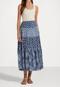 Blå mönstrad maxikjol med lagerdesign och blommotiven, parat med en krämfärgad kabelstickad linne. Har en åtsittande midja och en flödande silhuett.