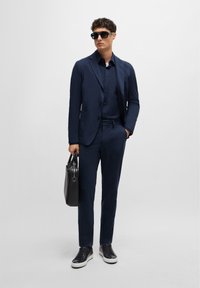 Navy suit with a slim fit, single-breasted jacket, matching shirt, black sunglasses, and black leather bag. Textured sneakers complete the look.