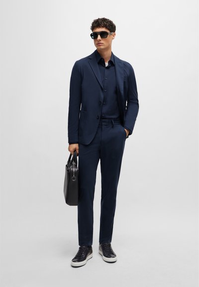 Costume marine ajusté, veste croisée simple, chemise assortie, lunettes de soleil noires et sac en cuir noir. Des baskets texturées complètent le look.
