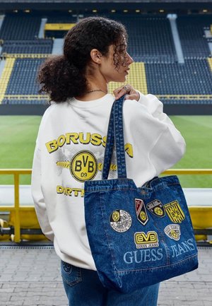 Junge Frau mit lockigem Haar hält eine Jeans-Tasche mit Borussia Dortmund-Aufnähern und steht in einem leeren Stadion, trägt einen weißen Borussia Dortmund-Pullover.