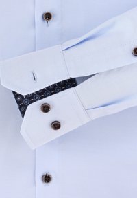 Světle modrá košile s rukávy, tmavohnědými knoflíky a ohnutým manžetovým krajkem, které odhaluje modro vzorovanou podšívku.