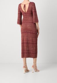 Robe rouge tricotée à manches trois-quarts, dos en V et détail de nœud. Le tissu présente un motif texturé et une silhouette ajustée jusqu'au mi-mollet.