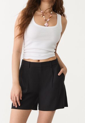 Frau trägt ein weißes, geripptes Tanktop, schwarze plissierte Shorts und eine Perlenkette mit pinken und braunen Akzenten.