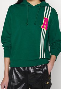 Felpa verde con cappuccio e lacci, con una lettera 'A' ricamata in rosa e tre strisce bianche sui lati. Il tessuto sembra morbido e testurizzato.