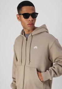 Bež zip-up dukserica s velikim prednjim džepovima, kapuljačom sa šniranjem i bijelim Nike logom. Nosena s crnim okruglim naočalama. Glatka tekstura tkanine.