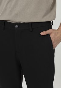 Zwarte geweven broeken met een vlakke voorkant, voorzien van een knoopsluiting en zijzakken, gecombineerd met een lichtbeige top.