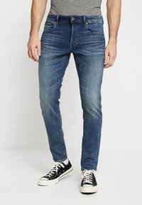 Herrjeans i blått denim med smal passform, lätt blekning, fem fickor och knappstängning, tillsammans med svarta sneakers.