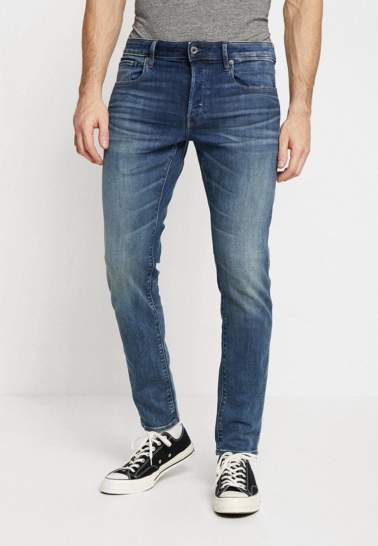 Herrjeans i blått denim med smal passform, lätt blekning, fem fickor och knappstängning, tillsammans med svarta sneakers.