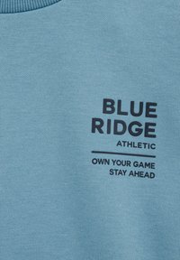 Svetlo modra športna majica s rebrastim okroglim izrezom. Značilno je črno besedilo: "BLUE RIDGE ATHLETIC" in "OWN YOUR GAME STAY AHEAD."