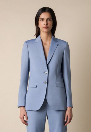 Frau mit glatten, dunklen Haaren trägt einen hellblauen, maßgeschneiderten Anzug mit zwei Knöpfen und Pattentaschen und steht vor einem beigen Hintergrund.
