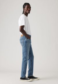 Jeans din denim deschis, cu un design drept, având buzunare la spate și o etichetă din piele maro. Asortați cu un tricou alb simplu și teniși negri.