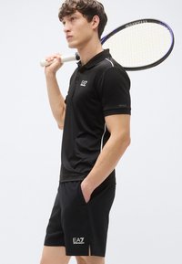 Juodas trumpomis rankovėmis polo marškinėlis su apykakle ir baltais akcentais, derinamas su juodais šortais, ant kurių yra logotipas ir šoninės skeltukai. Teniso raketė laikoma ant peties.