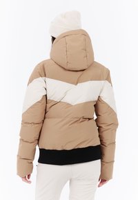 Nafúkaná bunda v béžovej a krémovej farbe s dizajnom v tvare šípok, so kapucňou a čiernym rebrovaným lemovaním na páse. Mäkký, izolovaný materiál.