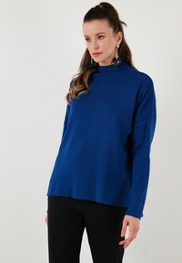 Pulover z dolgimi rokavi v modri barvi z visokim ovratnikom, sproščenega kroja in rebrasto teksturo, v paru s črnimi hlačami.